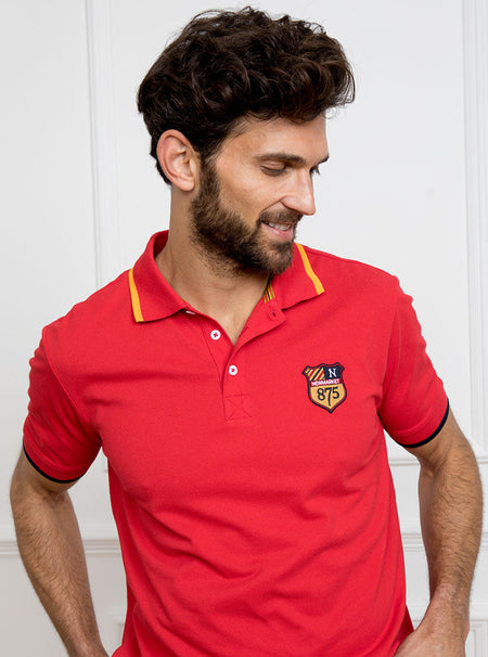 Man wearing red polo shirt in 100% cotton with Newmarket 875 crest embroidered on the front with navy sleeve trimming and gold collar trimming.